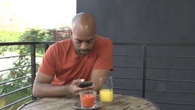 A mid-shot of a black man, using his cell phone, welcomes his partner, a white woman, to the breakfast table with a kiss. They sit and talk affectionately.