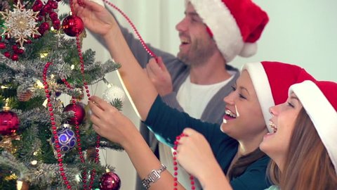 Young happy joyful family of a mother, father and teen daughter dressed in Santa Claus hat decorating Christmas and  New Year tree together at home, putting red ornaments. HD 1080p, Slow motion 240fps