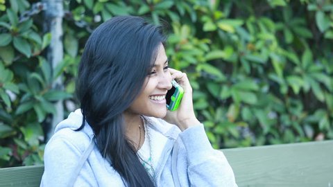 Closeup portrait, young, happy beautiful woman in gray coat sitting down outside in cold weather, speaking on cell phone, isolated background of green bushes