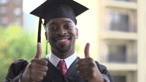 A close-up of a black man, dressed for graduation. He turns to camera and gives a thumbs-up of excitement.