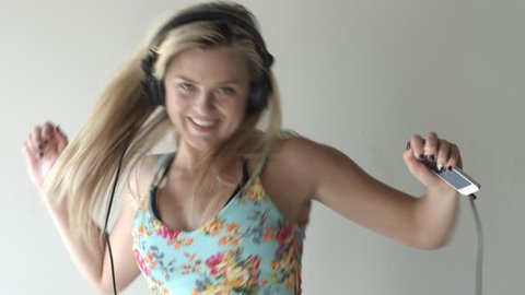Tilt up from feet to the face of a young blonde woman in blue dress, smiling and dancing while wearing headphones connected to a cellphone playing music.  Medium close up.  Recorded in 4K, UHD.