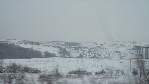 View captured on the window of a train over snowy fields, the site of a highway bridge, seen through the trees on the side of the railway.
