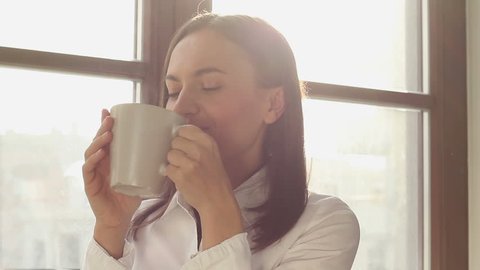 Beautiful young woman dreaming with cup of hot coffee over window