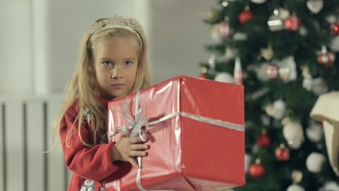 Cute little girl stands near stylishly decorated Christmas tree with a big gift box packed in red paper and examines a gift
