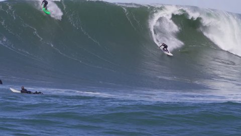 Two male surfers catch big wave at Mavericks, CA. Jet skis with filmers and safety team in foreground.