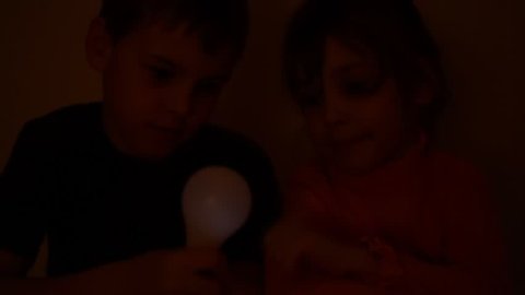 In the dark, a little girl in an orange blouse switches a light bulb on and off, which boy in blue T-shirt holds in his hand. They then raise the light bulb up.
