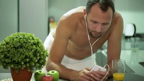 Man in towel watching video on smartphone and drinking juice after shower in kitchen
