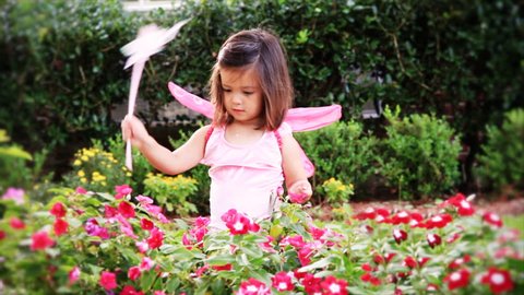 A darling little girl dressed in a fairy costume, tapes on a flowering plant with her pink wand.