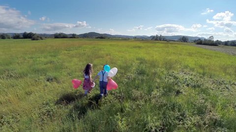 Two kids feel freedom and running with balloons. Aerial slow motion flaying by shoot of two kids, boy and girl, holding their hands running on a big grass field