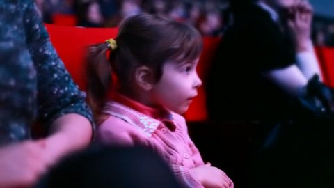 Impressionable little girl sits on a red chair in auditorium of circus, watches a show and claps