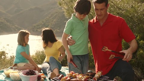 Dolly Shot Of Family Having Barbecue Picnic By Lake In Countryside.