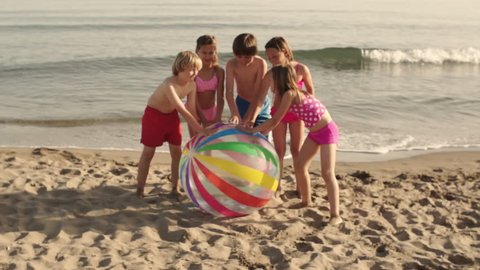 Slow-Motion Of Five Children Running Towards Camera Pushing Beach Ball On Beach.