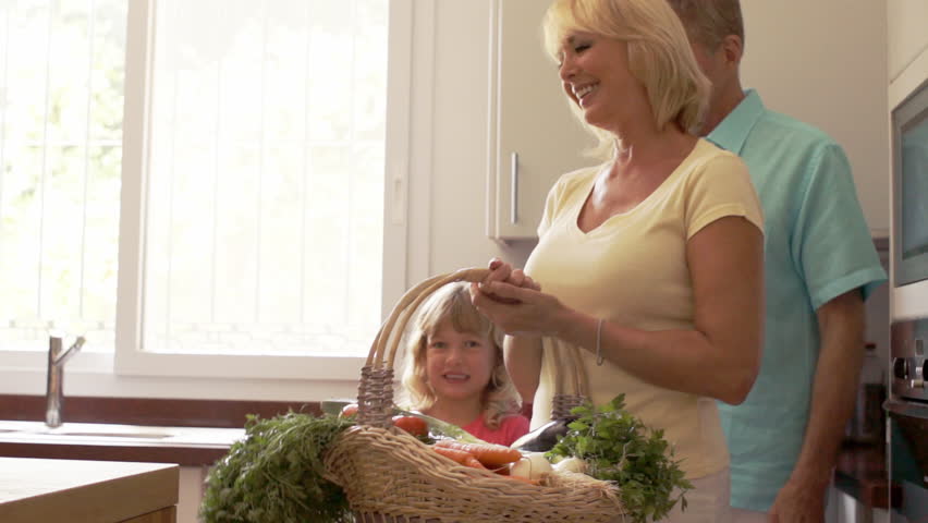 Grandparents And Granddaughter In Kitchen With Fresh Vegetables In Basket.