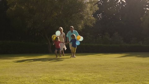 Slow-Motion Of Grandchildren And Grandparents Running To Camera In Garden Holding Balloons.