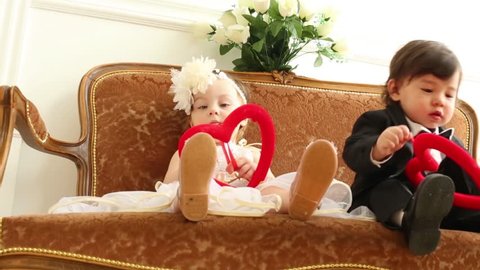 Babies in costumes of bride and groom with red hearts sit on couch. Boy peels off