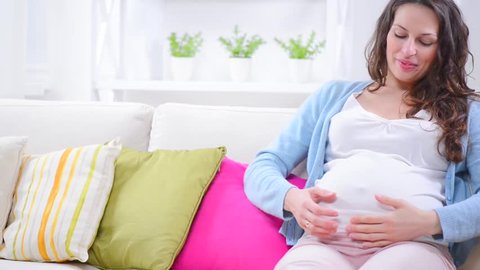 Happy pregnant woman resting on a sofa and caressing her tummy. Young relaxed pregnant woman touching her belly sitting on couch at home. Expectant young woman HD 1080p, Dolly shot