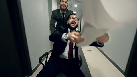 Guys celebrating a deal riding a chair in the office hall and tossing documents