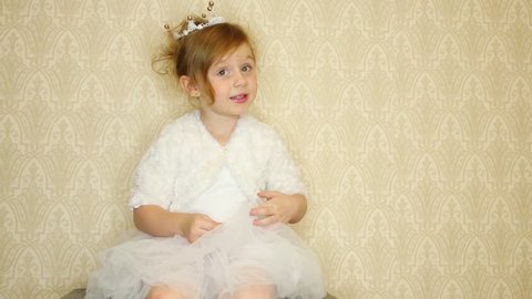 Little girl wearing a crown and white dress sitting against the wall and conjures with magic wand