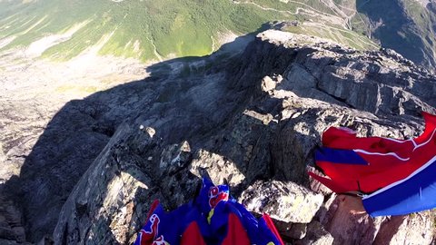 Two skydivers in wingsuits jump from a cliff before gliding in the air over a green mountain landscape, POV