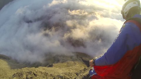 A base jumper in a wingsuit jumps down from a cliff, passes through clouds while gliding over a landscape, POV
