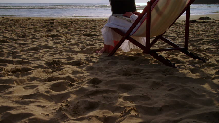 A middle aged woman sits in an armchair on the beach and uses her laptop.
