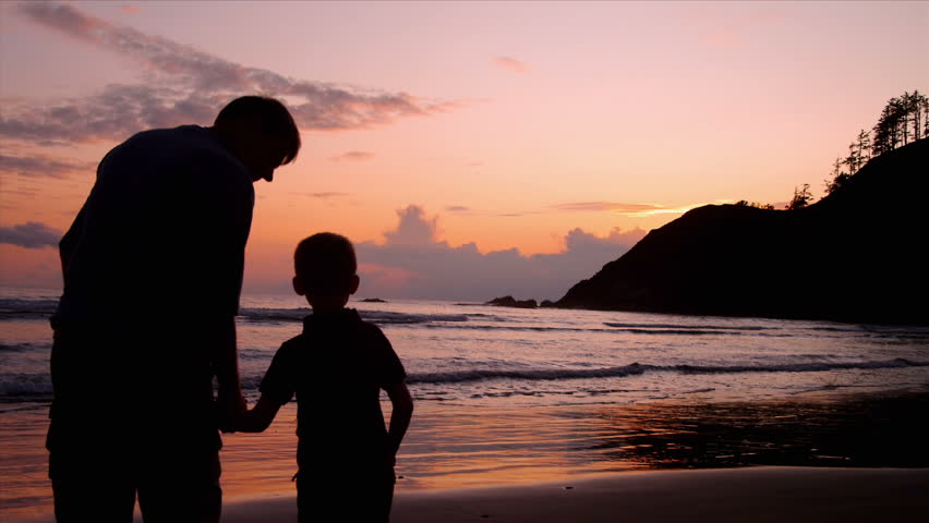 Father and son hold hands on a beach at sunset.