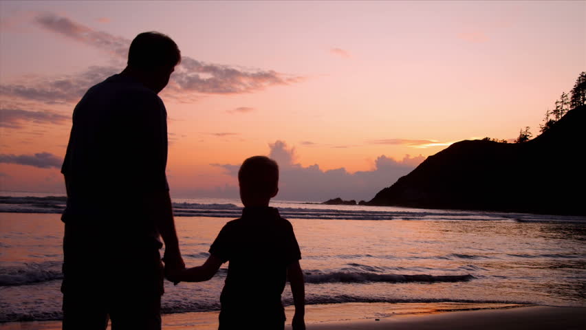 Father and son hold hands on a beach at sunset.