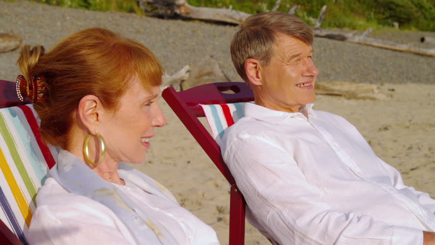 An elderly couple sits on the beach together and holds hands