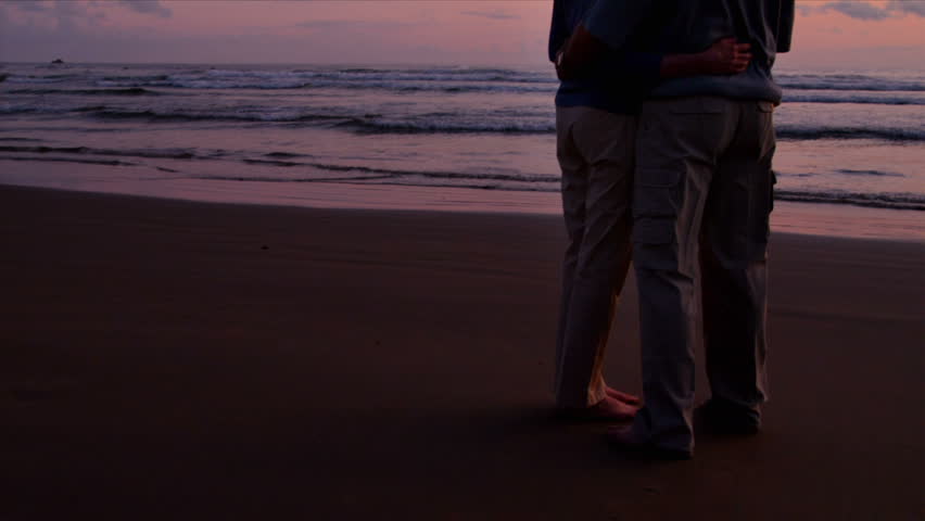 An elderly couple embraces on the beach at sunset. 