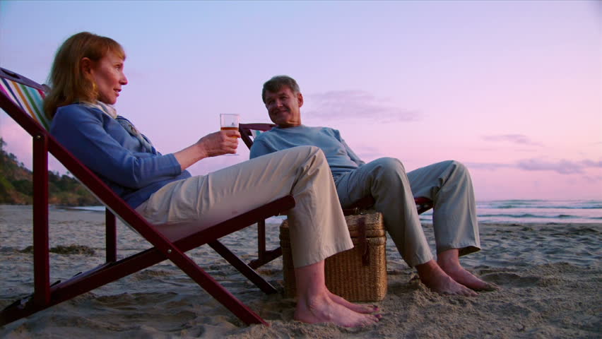 An elderly couple toasts glasses on the beach at sunset. 