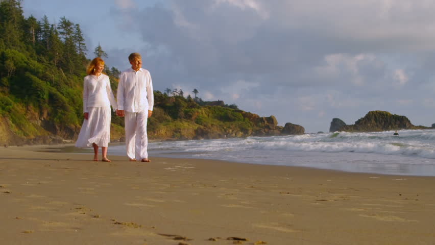 Older couple hold hands and walk on the beach together. 