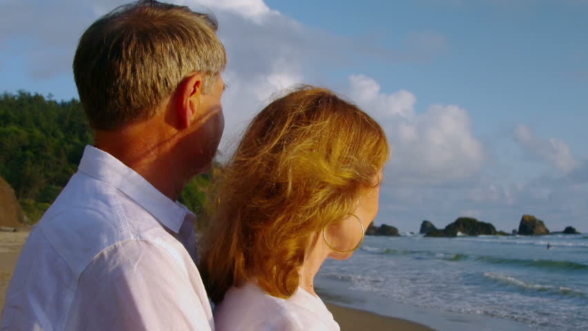 Older couple embrace on the beach while looking at the water. 