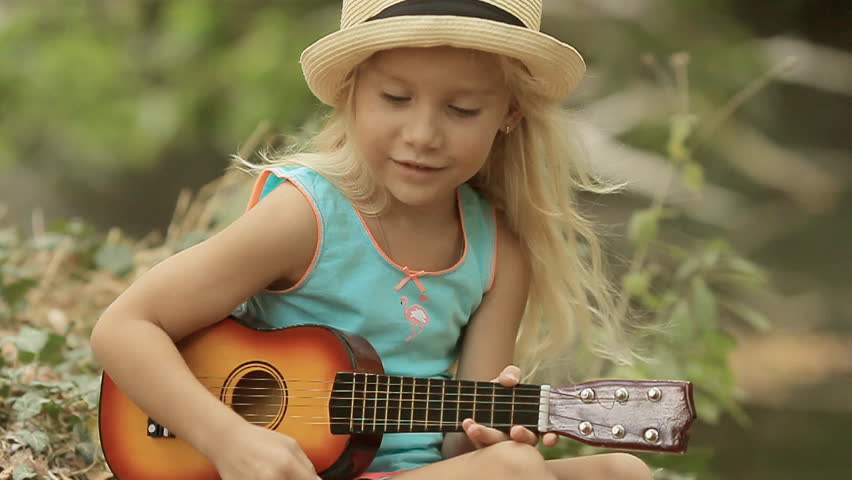 Beautiful little girl in straw hat playing on toy guitar in the woods next to the river