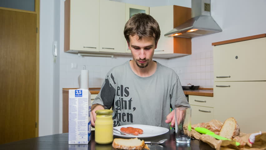 Man in dining room cutting hot-dogs. Wide shot of person behind kitchen table eating fried hot-dogs, with mustard and milk beside.