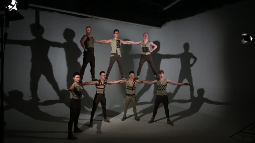 Seven guys in medieval costumes do acrobatic pyramid in grey studio with lamps