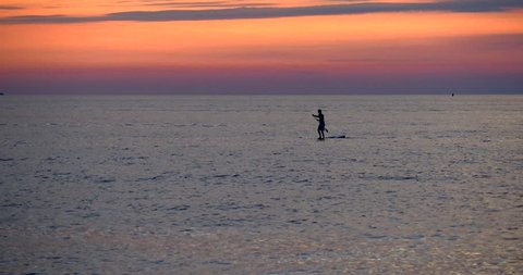 Dark silhouette of a surfer man person standing up on a paddle board (SUP) or surfboard and paddleboarding on the sea water of Pacific ocean near Hawaii beach at sunset in summer. Sport, lifestyle 4K.