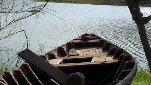 Viewing down the length of an old wooden boat resting on the banks of a lake. The surface of the lake is disturbed by a gentle breeze. A small flock of tropical birds pass from left to right.