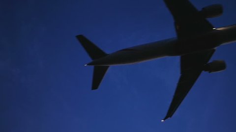 Low angle slow motion view of an airplane silhouette flying overhead flashing red lights in dark sky. Modern twin jet about to land at night.