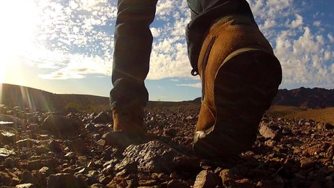 Low and wide angle shot of a man as he walks away from camera in a barren rocky desert. A hiker enjoys exploring a rough scenic landscape strewn about with small stones.