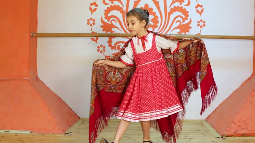 Smiling girl in red folk costume dances and spins with shawl at stage.