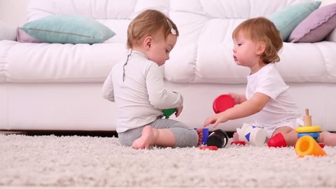 Two pretty girls sit on soft carpet and play with toys near white sofa