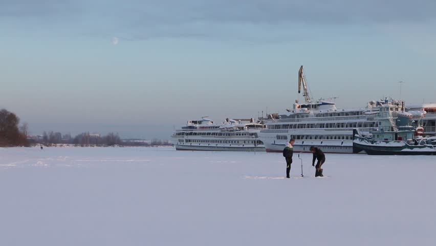 Two fishermen catch fish on frozen river near tourist ship at winter day
