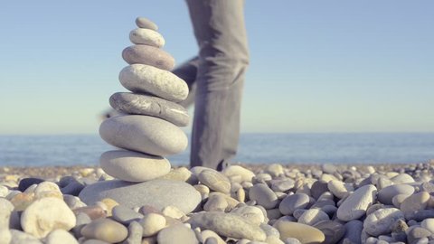 Stack of pebbles balancing one on another, making a tower. Close-up shot on pebble beach with the sea/ocean in the background; a man walks by; sunny day. Very Zen.
