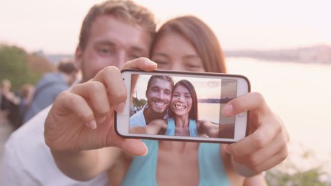 Smart phone selfie - couple taking self portrait using smartphone camera. Dating couple in love having fun taking candid fresh picture photo laughing smiling. Caucasian man, Asian woman at sunset.