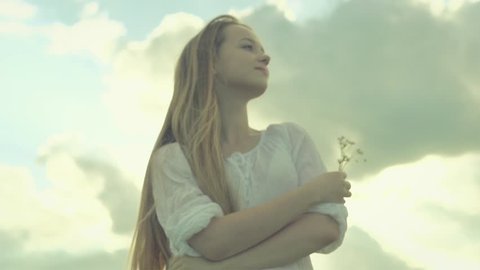 Beauty model girl relaxing on spring field. Beautiful young woman over cloudy sky enjoying nature and smelling wild flowers. Slow motion video footage full HD 1080. High speed camera shot 240 fps
