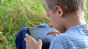 portrait of boy playing computer games on the tablet pc outdoors in park. children spare-time. kid using tablet computer touchscreen. child sitting alone in green grass. modern toys. close up. hd
