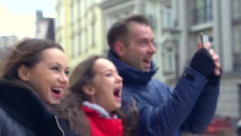 Young happy tourist family having tour walking in city. Amazed Girl showing landmarks. Enjoying sights of town. Man taking selfie photo outside. Tourism concept. Slowmotion 240 fps, 1080p
