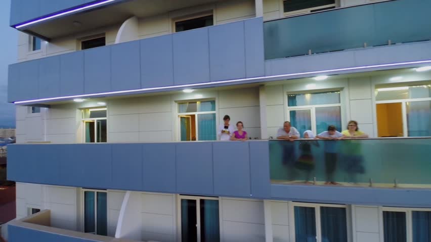 Six people of three family generations stand on balcony of hotel at summer evening. Aerial view