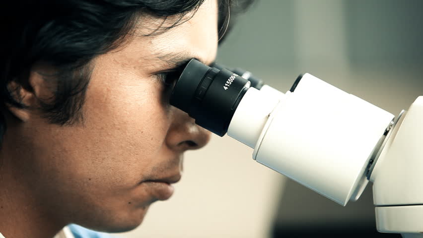 Close up of a scientist who is looking through a microscope while researching /