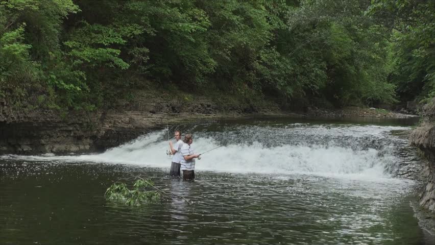 Two fishermen in white t-shirt and shorts on small river. One man catch trout under waterfall. The film was shot in late May in sunny day.